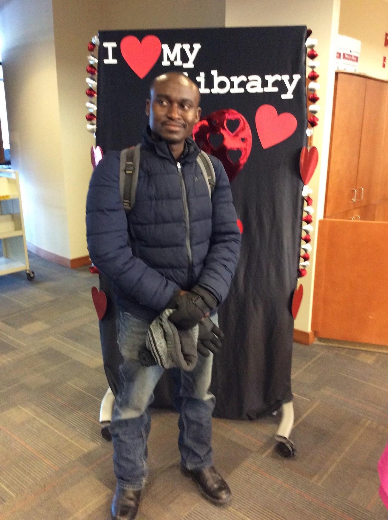 A student in winter gear stands in front of the I love my library backdrop, looking off to one side.
